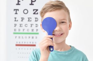Child standing in front of eye exam chart smiles while covering one eye with an eye occluder