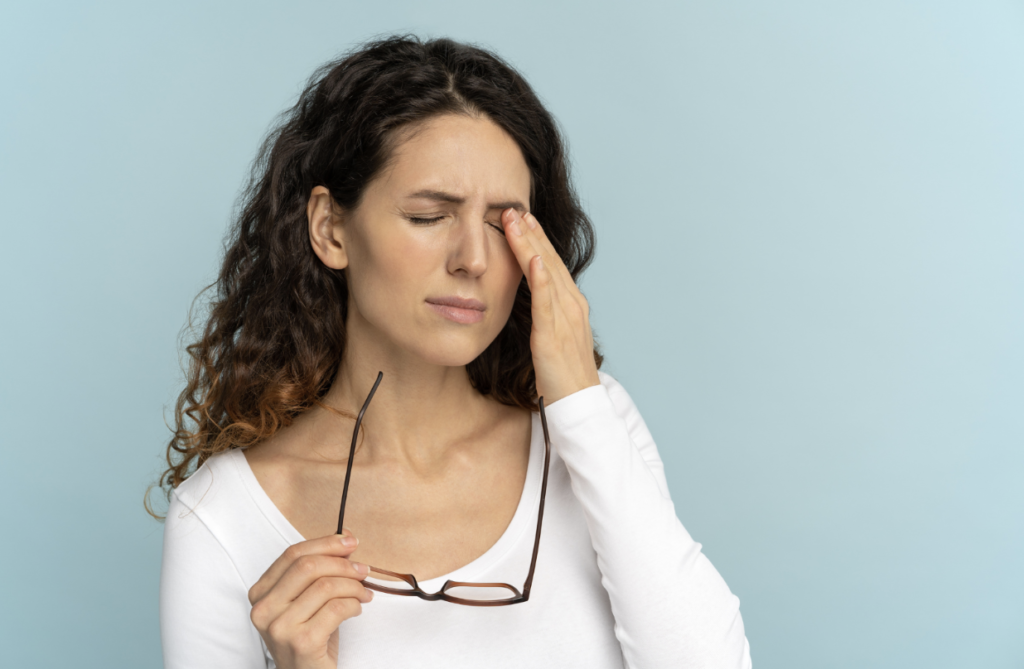 A woman taking off her glasses, rubbing her eyes while squinting due to eye pain