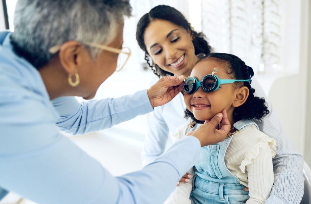 A smiling child sits in their mother's lap for an eye exam while their optometrist fits them for glasses.